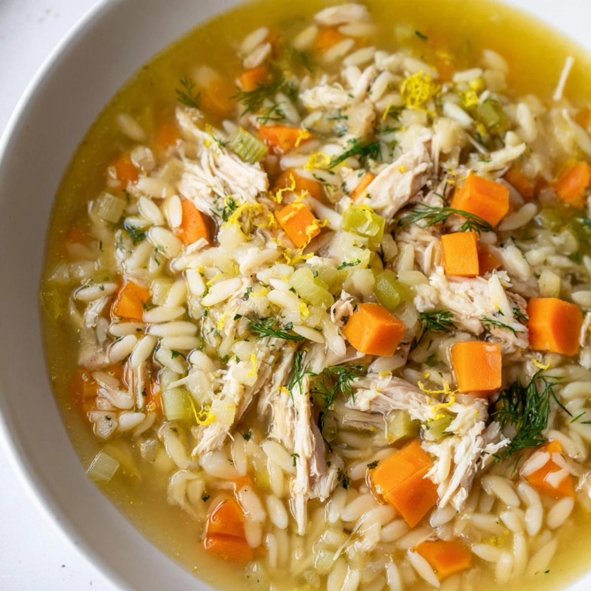 A steaming bowl of Chicken Lemon Orzo Soup garnished with fresh dill and lemon zest, served hot in a rustic ceramic bowl.  